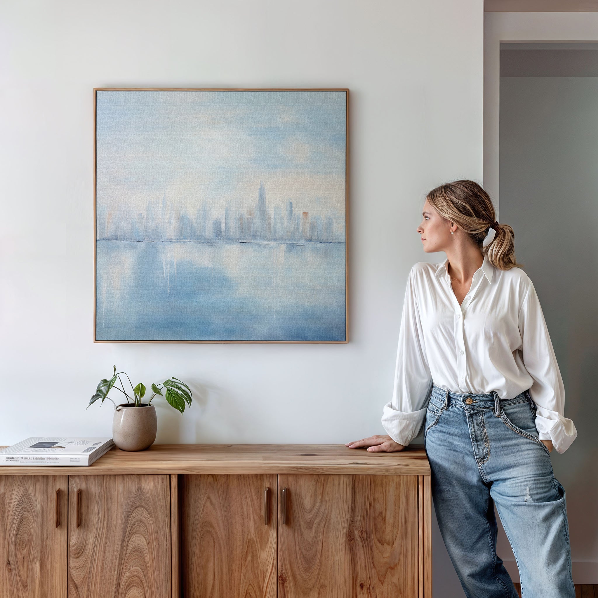 A woman in a white blouse and jeans leans on a wooden cabinet, admiring the Urban Mirage Canvas Art - Square with its dreamy city skyline. A potted plant and books complete the stylish modern living room decor.