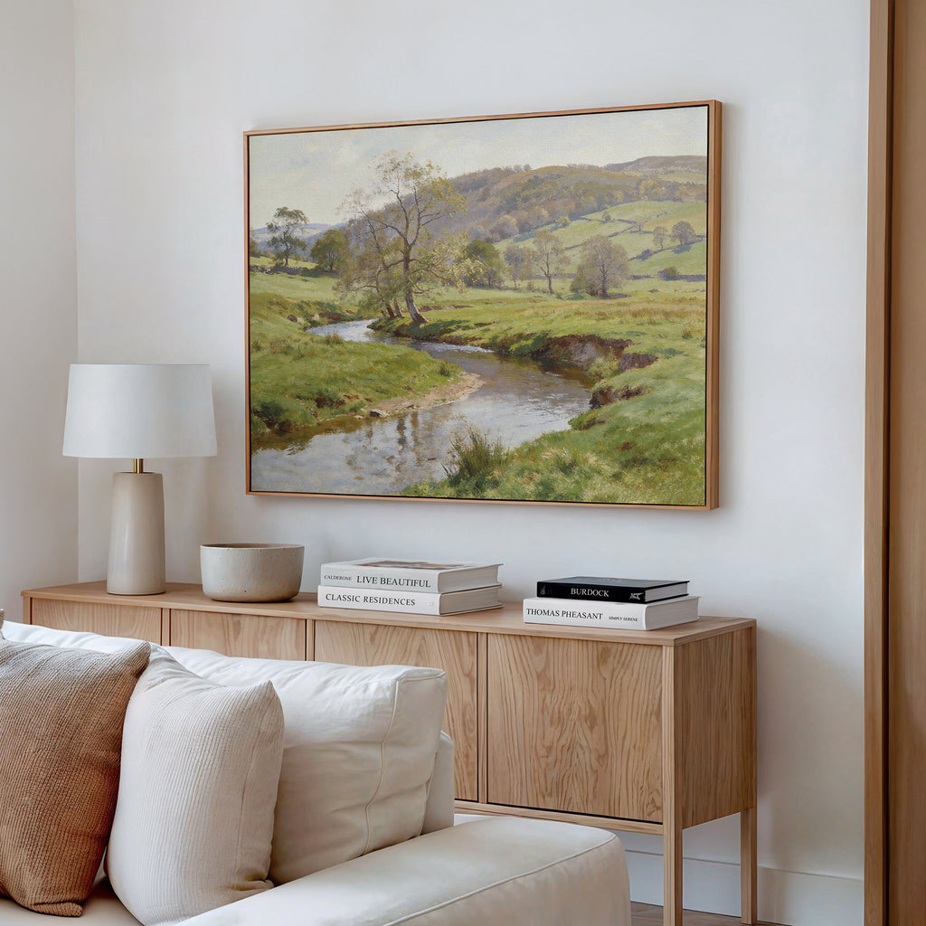 A cozy living room with a light wood sideboard, white table lamp, stacked books, decorative bowls, and the Valley Stream Canvas Art - Horizontal featuring a countryside river and trees hanging on the wall above.