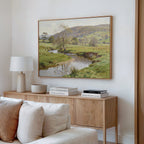 A cozy living room with a light wood sideboard, white table lamp, stacked books, decorative bowls, and the Valley Stream Canvas Art - Horizontal featuring a countryside river and trees hanging on the wall above.