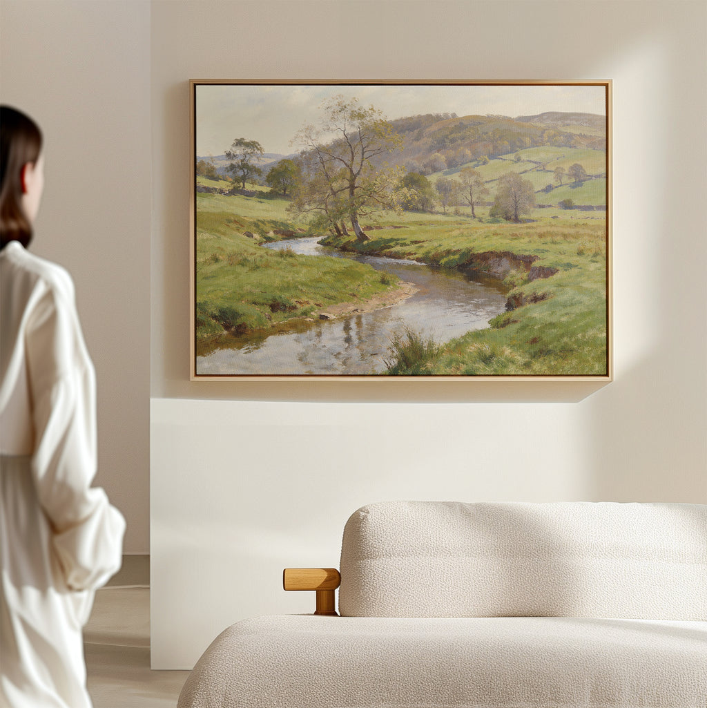 A woman stands in a softly lit room, admiring the Valley Stream Canvas Art - Horizontal above a beige sofa, featuring a river winding through lush green countryside.