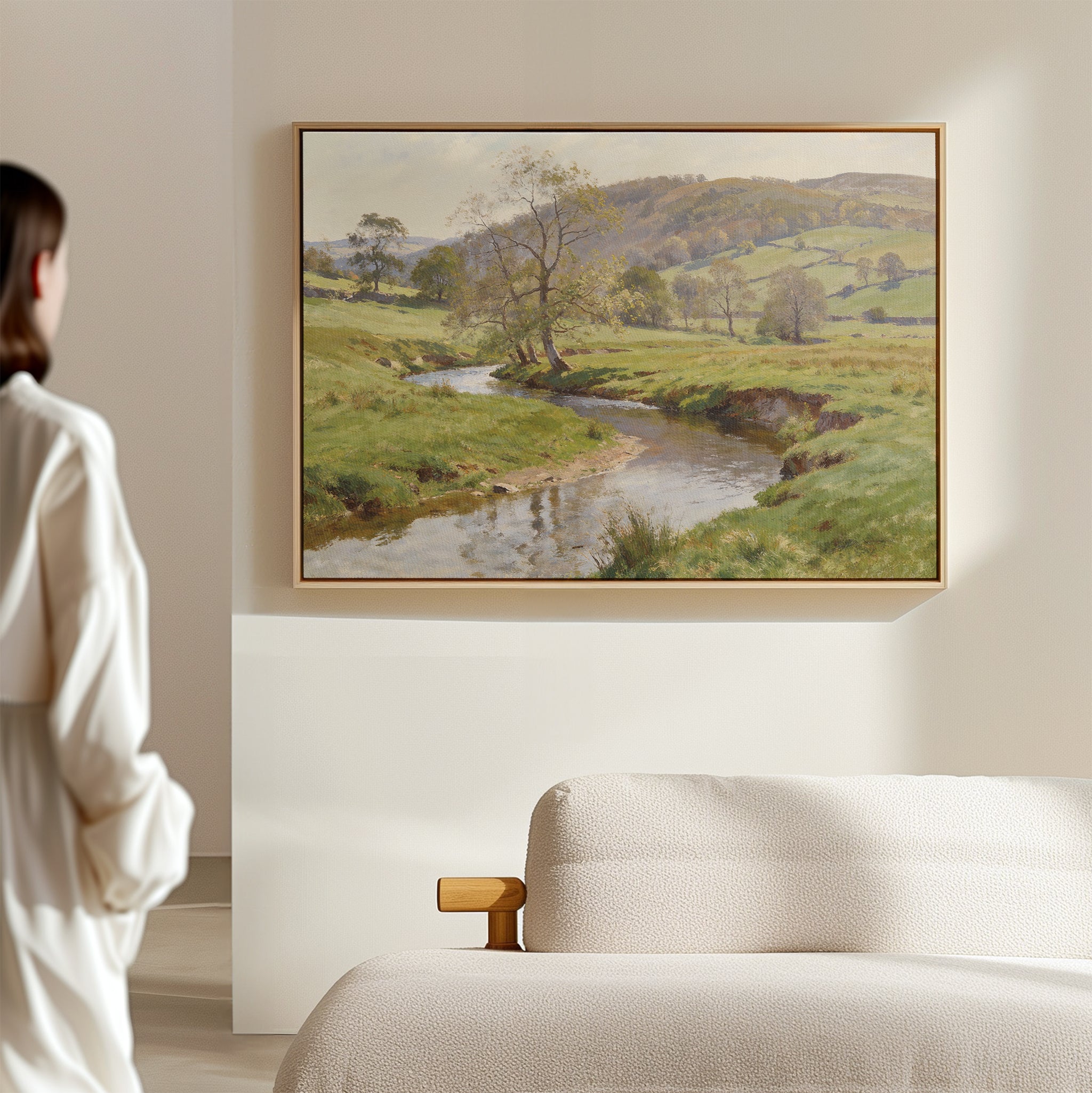 A woman stands in a softly lit room, admiring the Valley Stream Canvas Art - Horizontal above a beige sofa, featuring a river winding through lush green countryside.