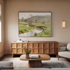 A cozy living room features a textured wooden sideboard, neutral sofas, a low wooden coffee table with a book and vase, and the Valley Stream Canvas Art - Horizontal above the sideboard, highlighting natural countryside beauty.