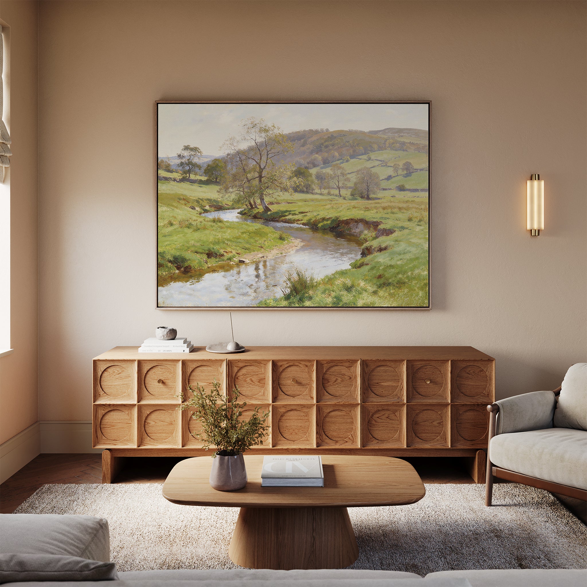 A cozy living room features a textured wooden sideboard, neutral sofas, a low wooden coffee table with a book and vase, and the Valley Stream Canvas Art - Horizontal above the sideboard, highlighting natural countryside beauty.