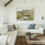 A cozy living room with a white sectional, assorted throw pillows, a wooden coffee table with decor, and the Valley Stream Canvas Art - Horizontal featuring a scenic stream and hills on a white shiplap wall.