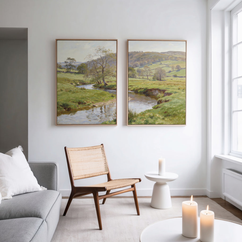 A bright, minimalist living room with a gray sofa, woven chair, and small white side table topped with a lit candle. Above the chair hang Valley Stream - Set of 2 wall art canvases of green countryside, while large windows bring in natural light.