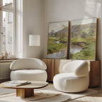 A bright, modern living room with two Valley Stream - Set of 2 curved white armchairs, a low wooden coffee table, tall floor lamp, and serene countryside canvas art leaning against the sideboard creates a peaceful ambiance.