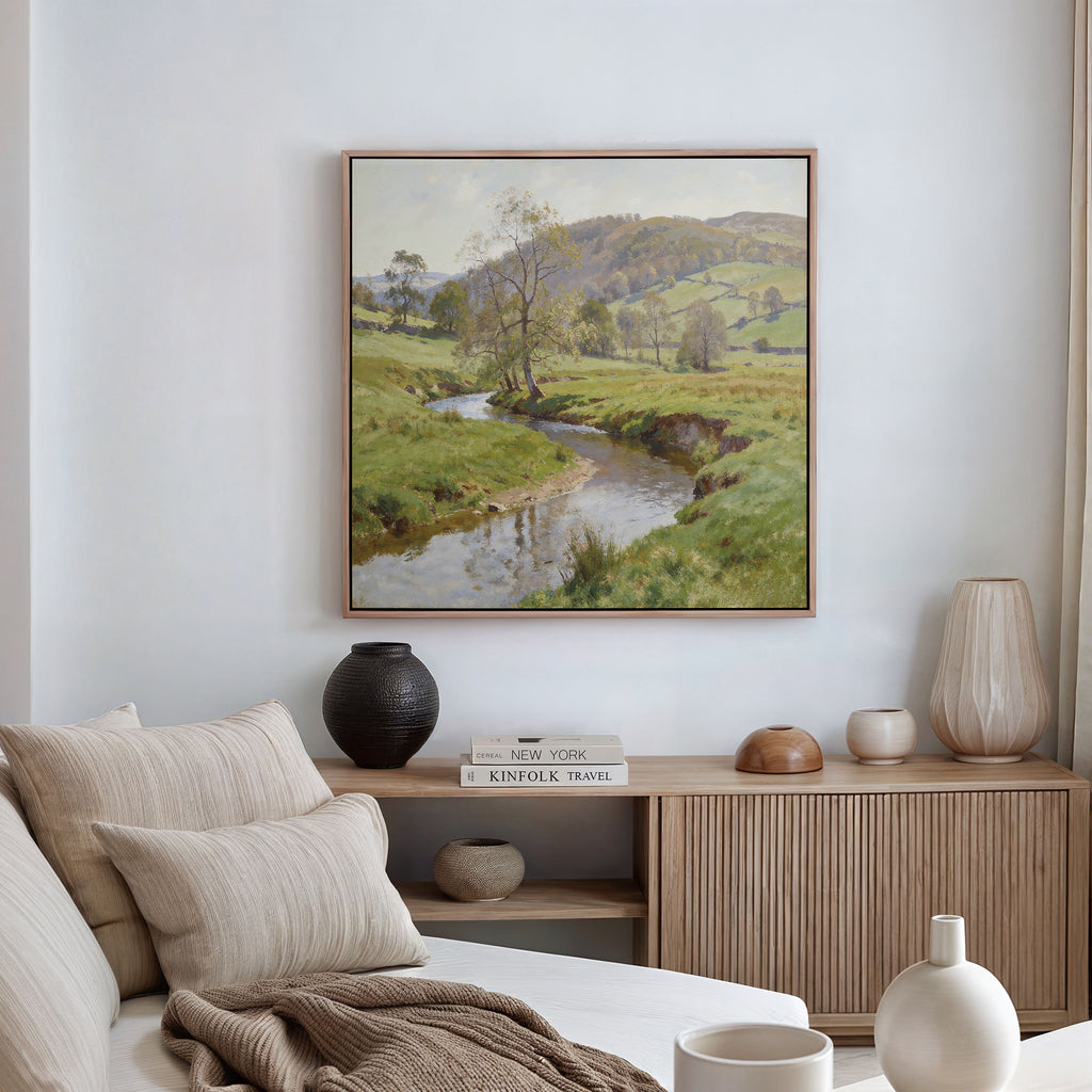 A cozy living room with a beige sofa, neutral pillows, wooden sideboard, decorative vases, and the Valley Stream Canvas Art - Square depicting a green countryside with a stream hanging above.