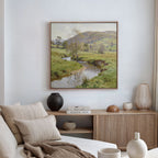 A cozy living room with a beige sofa, neutral pillows, wooden sideboard, decorative vases, and the Valley Stream Canvas Art - Square depicting a green countryside with a stream hanging above.