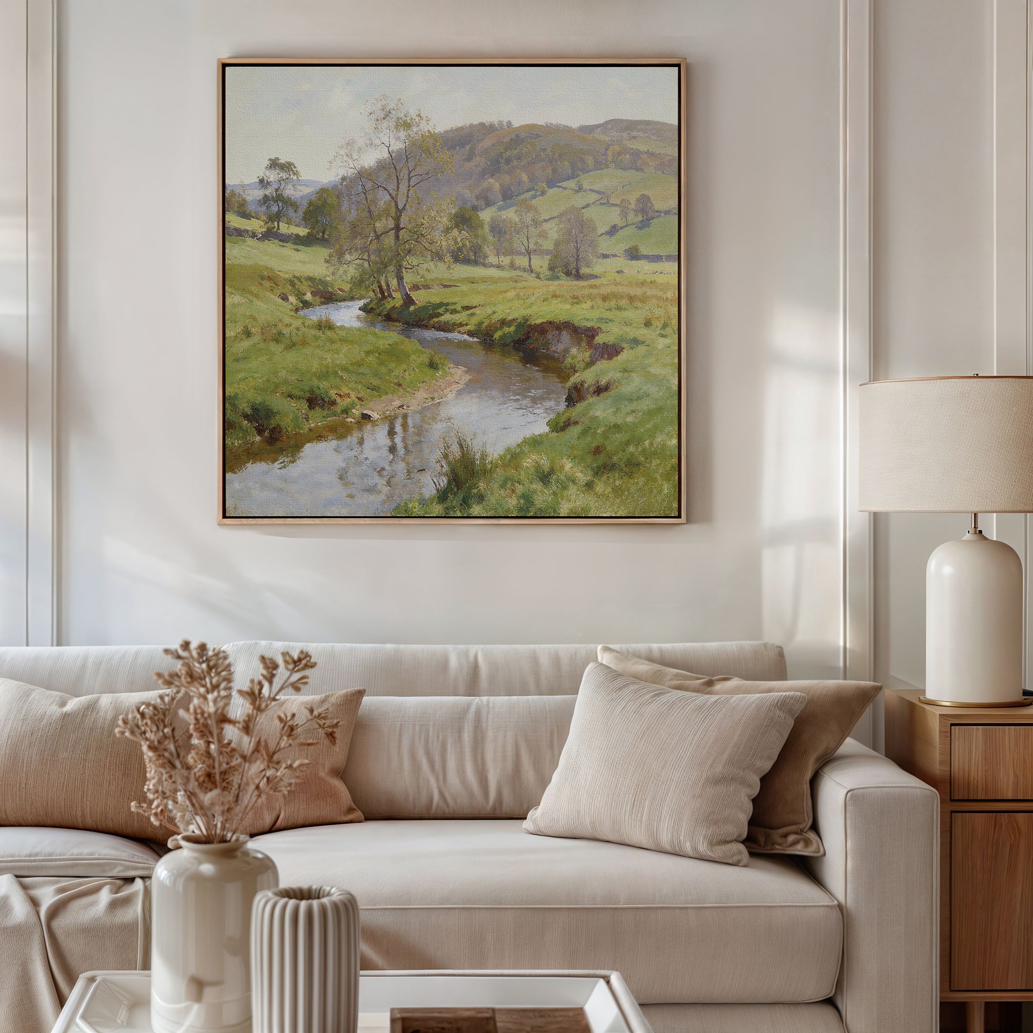 A living room with a beige sofa, cream cushions, a side table with lamp, and dried flowers. Above the sofa is the Valley Stream Canvas Art - Square, its serene river and hills bringing calm Nordic Park wall art inspiration to the space.