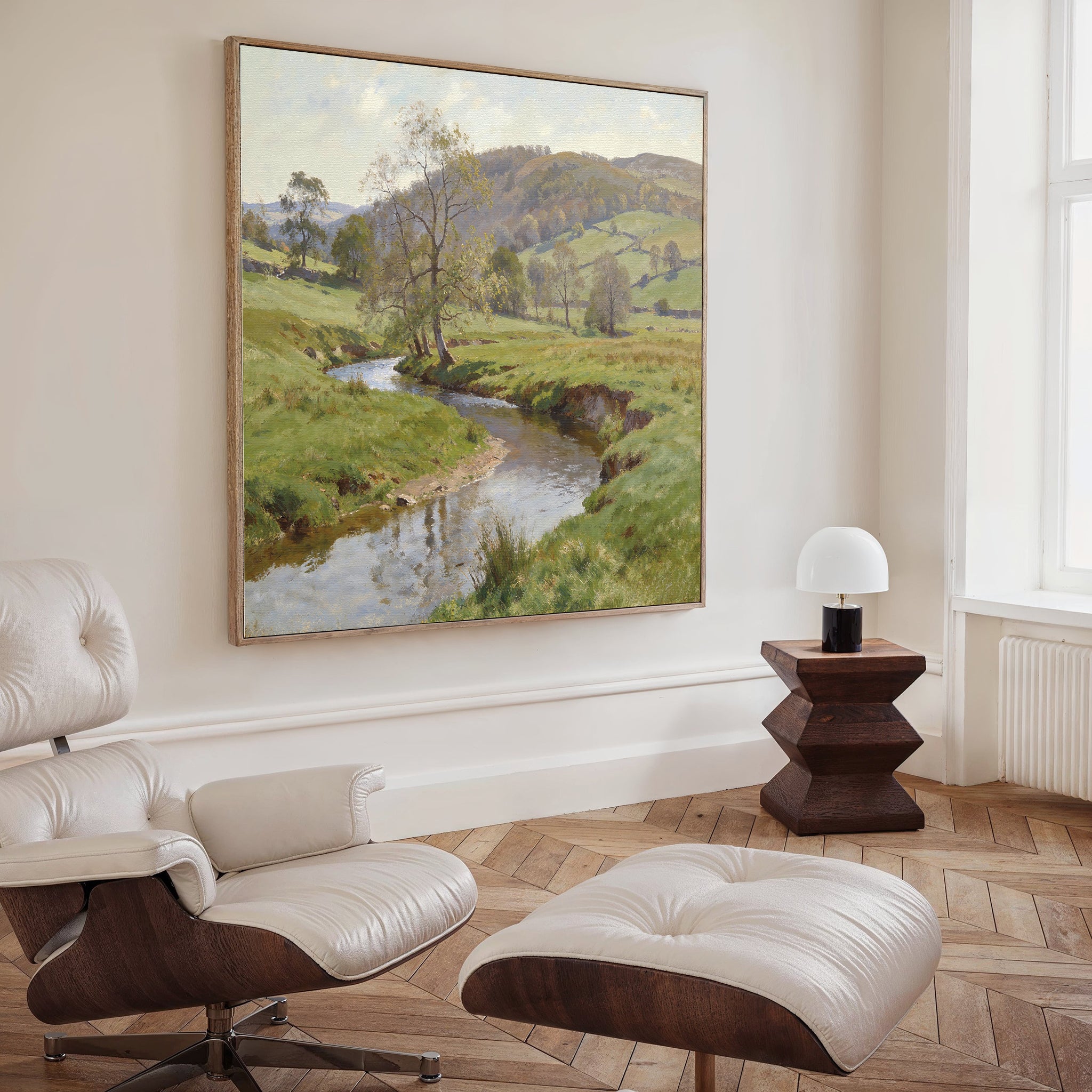 A modern living room with a white leather lounge chair and ottoman, a small dark wood side table with a white lamp, and the Valley Stream Canvas Art - Square depicting a river and green hills hanging on the wall.