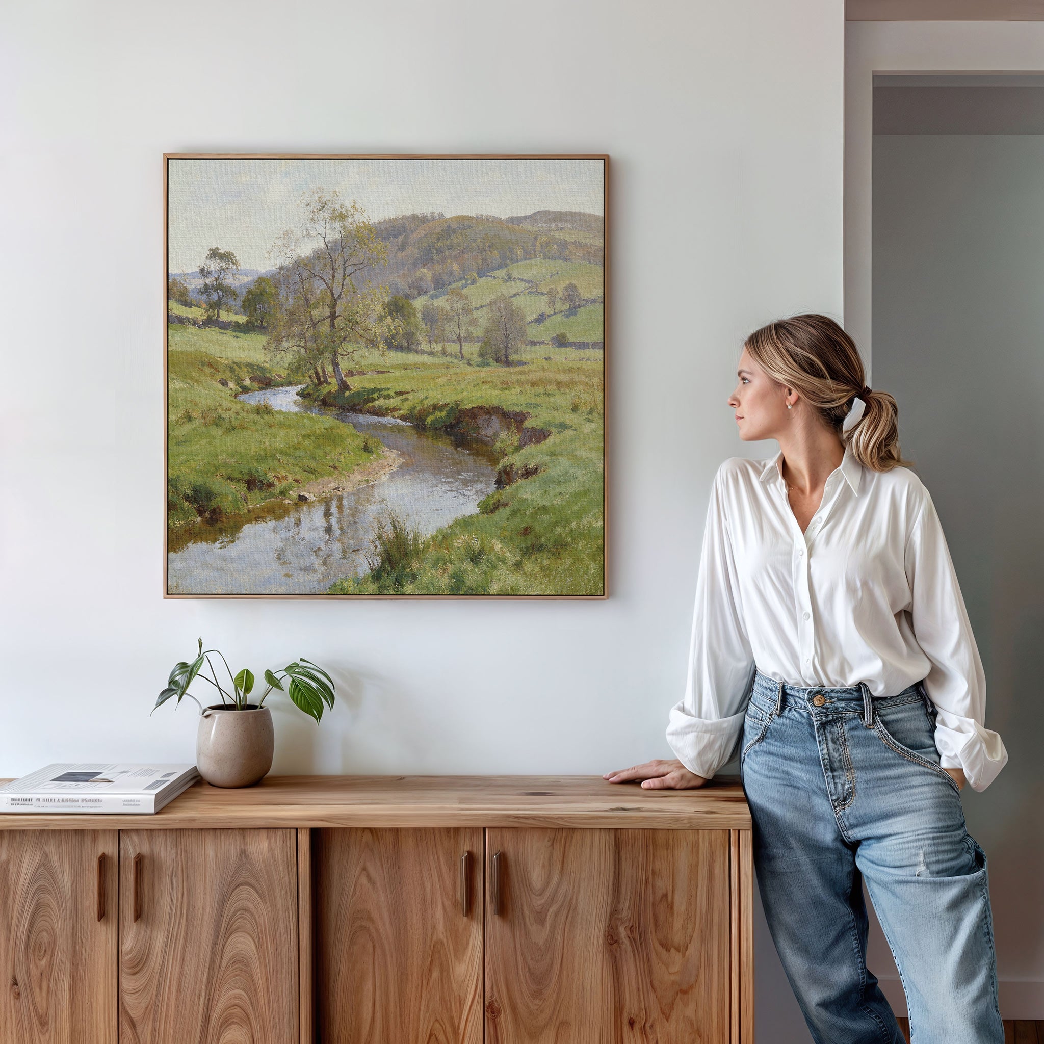 A woman in a white blouse and jeans leans on a wooden cabinet, gazing at the Valley Stream Canvas Art - Square featuring green countryside with a stream. A potted plant and book rest on the cabinet below the wall art.