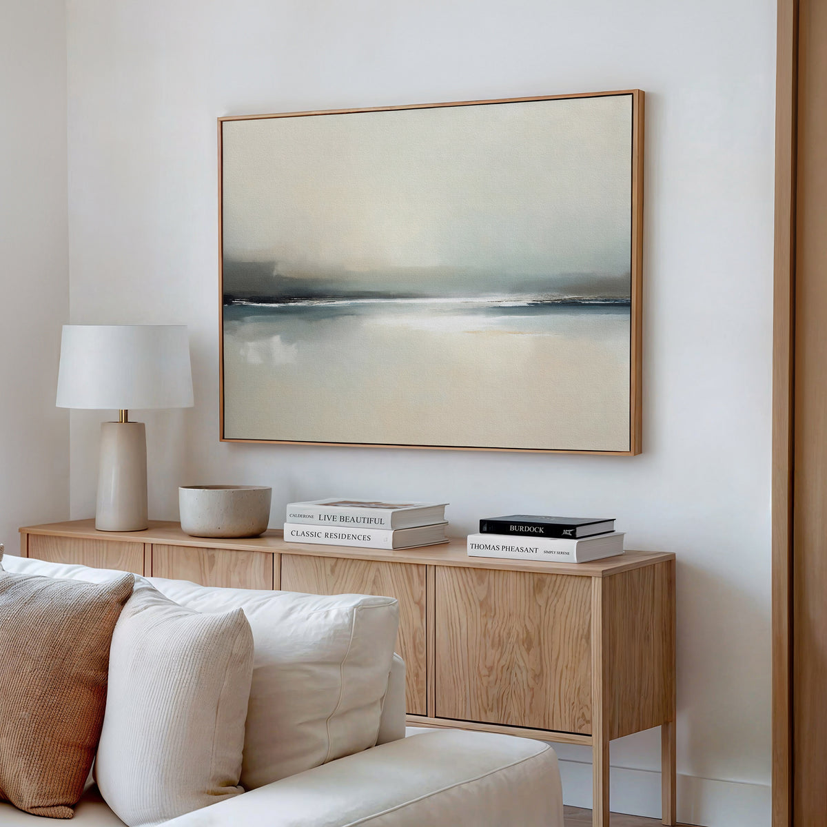 A modern living room with a light wood sideboard, stacked books, a white lamp, a decorative bowl, and the Veiled Estuary Canvas Art – Horizontal in soft neutral tones above a cream-colored sofa on a white wall.