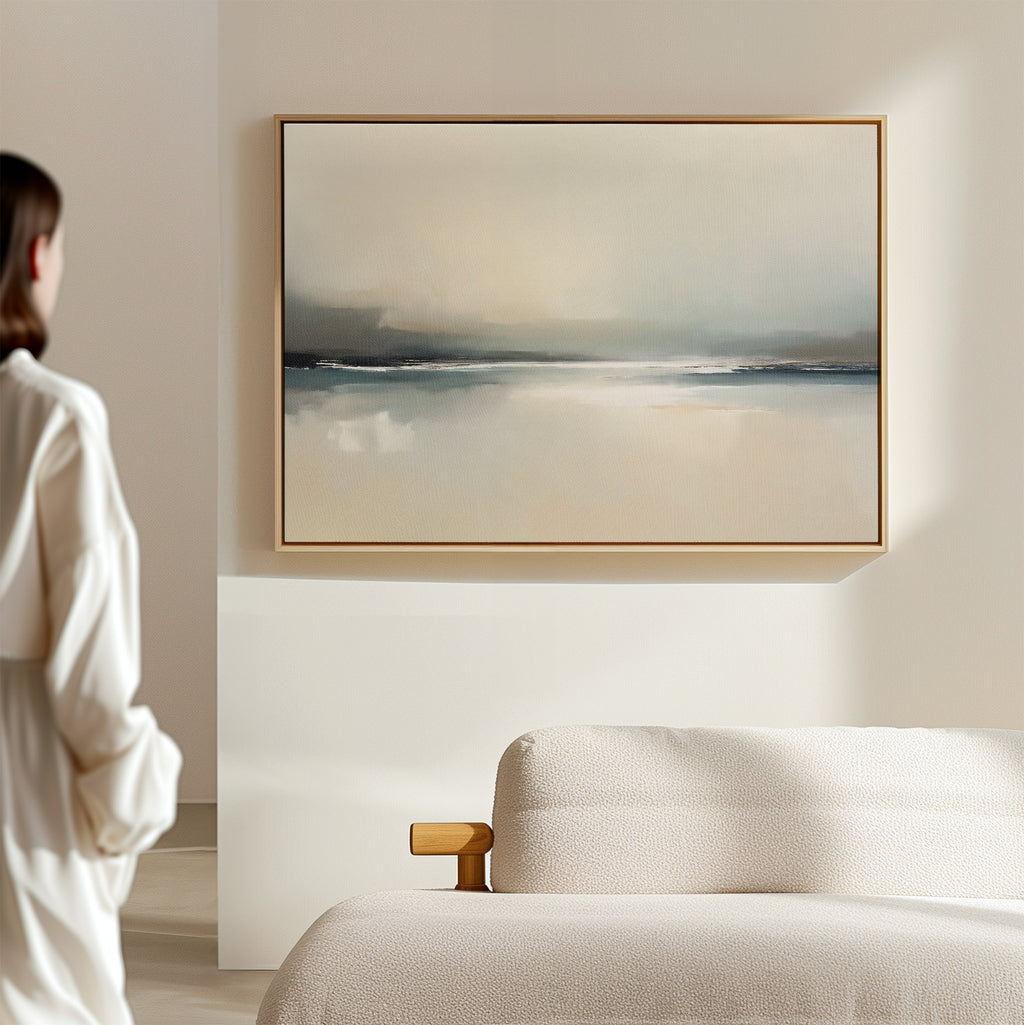 A person in a white shirt stands in a minimalist room, gazing at the Veiled Estuary Canvas Art - Horizontal with soft blue and beige tones displayed above a beige sofa, creating a calm and serene atmosphere.