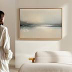 A person in a white shirt stands in a minimalist room, gazing at the Veiled Estuary Canvas Art - Horizontal with soft blue and beige tones displayed above a beige sofa, creating a calm and serene atmosphere.