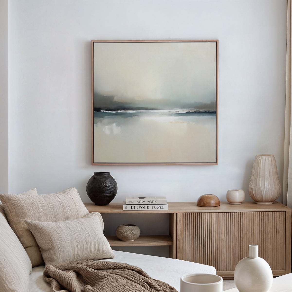 A modern living room with a neutral palette features a beige sofa, wooden sideboard with vases and books, and the Veiled Estuary Canvas Art - Square—a large abstract painting in soft blue, gray, and beige—displayed on the wall.