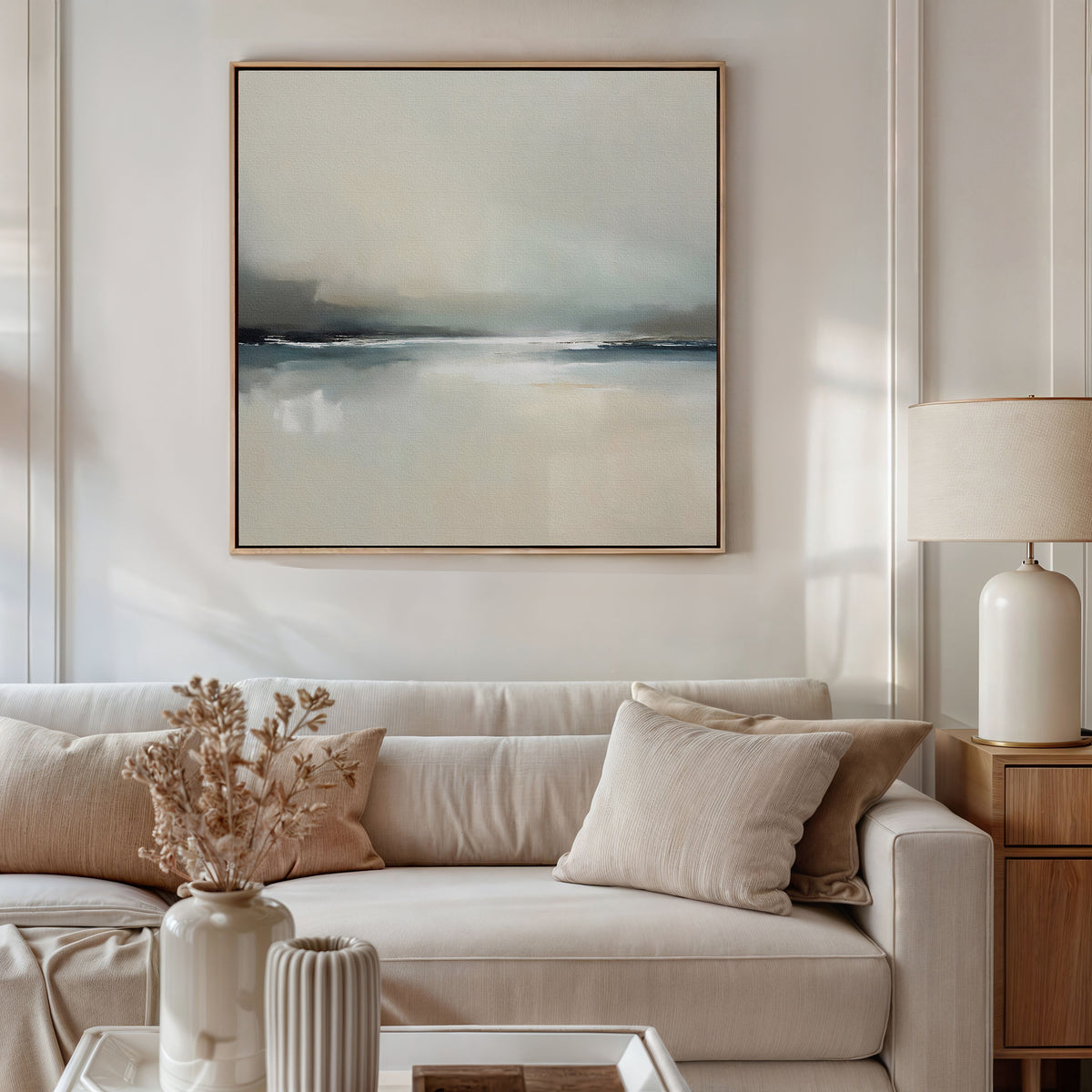A modern living room with a beige sofa, neutral cushions, side table and ceramic lamp, vase of dried flowers, and the Veiled Estuary Canvas Art - Square featuring a serene landscape above the sofa.