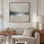 A modern living room with a beige sofa, neutral cushions, side table and ceramic lamp, vase of dried flowers, and the Veiled Estuary Canvas Art - Square featuring a serene landscape above the sofa.