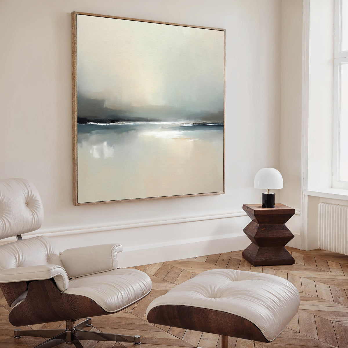 A modern, minimalist living room with a white lounge chair and ottoman, a geometric wooden side table with a small lamp, and Veiled Estuary Canvas Art - Square displaying soft, muted colors on the wall.