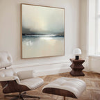 A modern, minimalist living room with a white lounge chair and ottoman, a geometric wooden side table with a small lamp, and Veiled Estuary Canvas Art - Square displaying soft, muted colors on the wall.