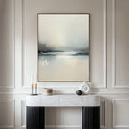 A modern console table displays decorative objects against a wall with elegant molding. Above hangs the Veiled Estuary Canvas Art - Vertical, its soft white, gray, and blue tones evoking the tranquil beauty of a Nordic landscape.