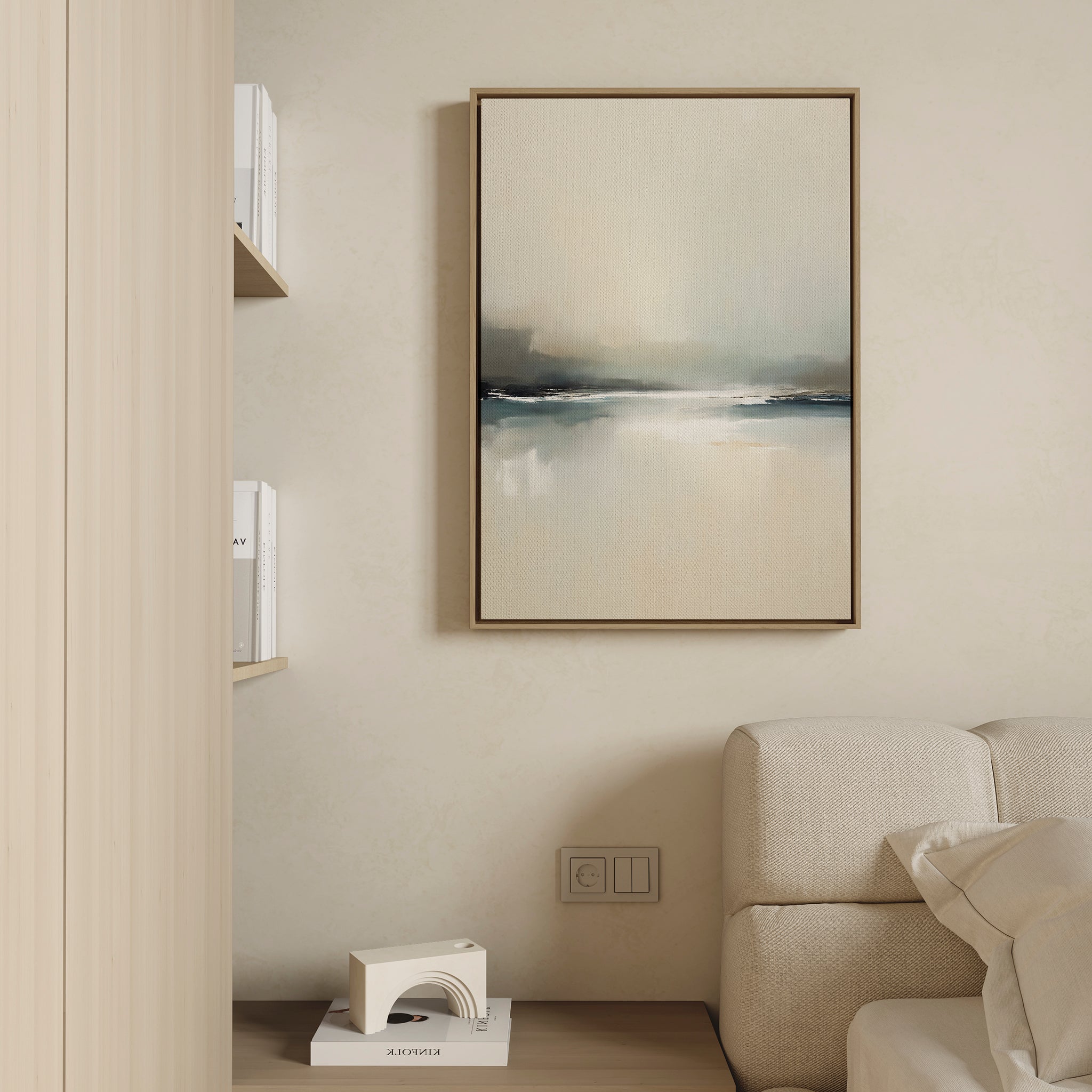 A minimalist living space features a beige sofa, light wood shelves with white books, and Veiled Estuary Canvas Art - Vertical—an abstract painting in soft blue, gray, and beige—enhancing the cream-colored wall.