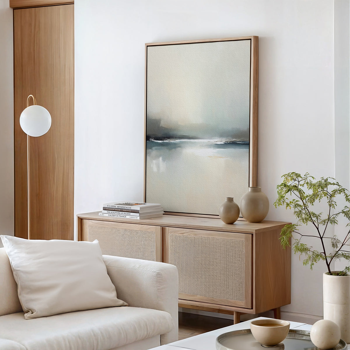 A modern living room features a light wooden sideboard with vases, books, and the Veiled Estuary Canvas Art - Vertical above it. A white sofa with cushions and a small indoor plant complete the neutral, calming decor.