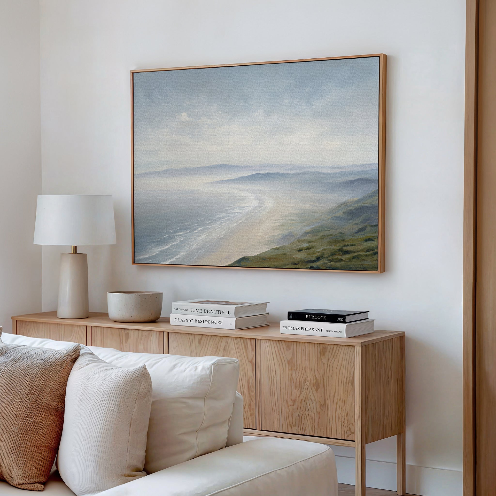 A minimalist living room featuring a wooden sideboard, books, a white lamp, and the Veiled Shores Canvas Art - Horizontal above, creating serene coastal Nordic Park vibes.
