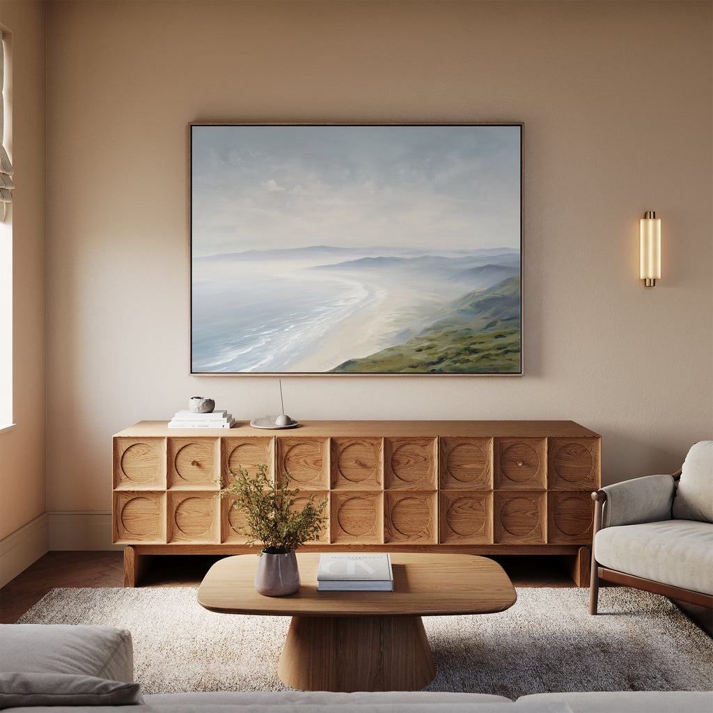 A cozy living room features a light wood sideboard, coffee table, and beige sofa. Above the sideboard hangs the Veiled Shores Canvas Art - Horizontal, depicting a serene coastal scene. A small lamp and vase add to the Nordic Park-inspired decor.