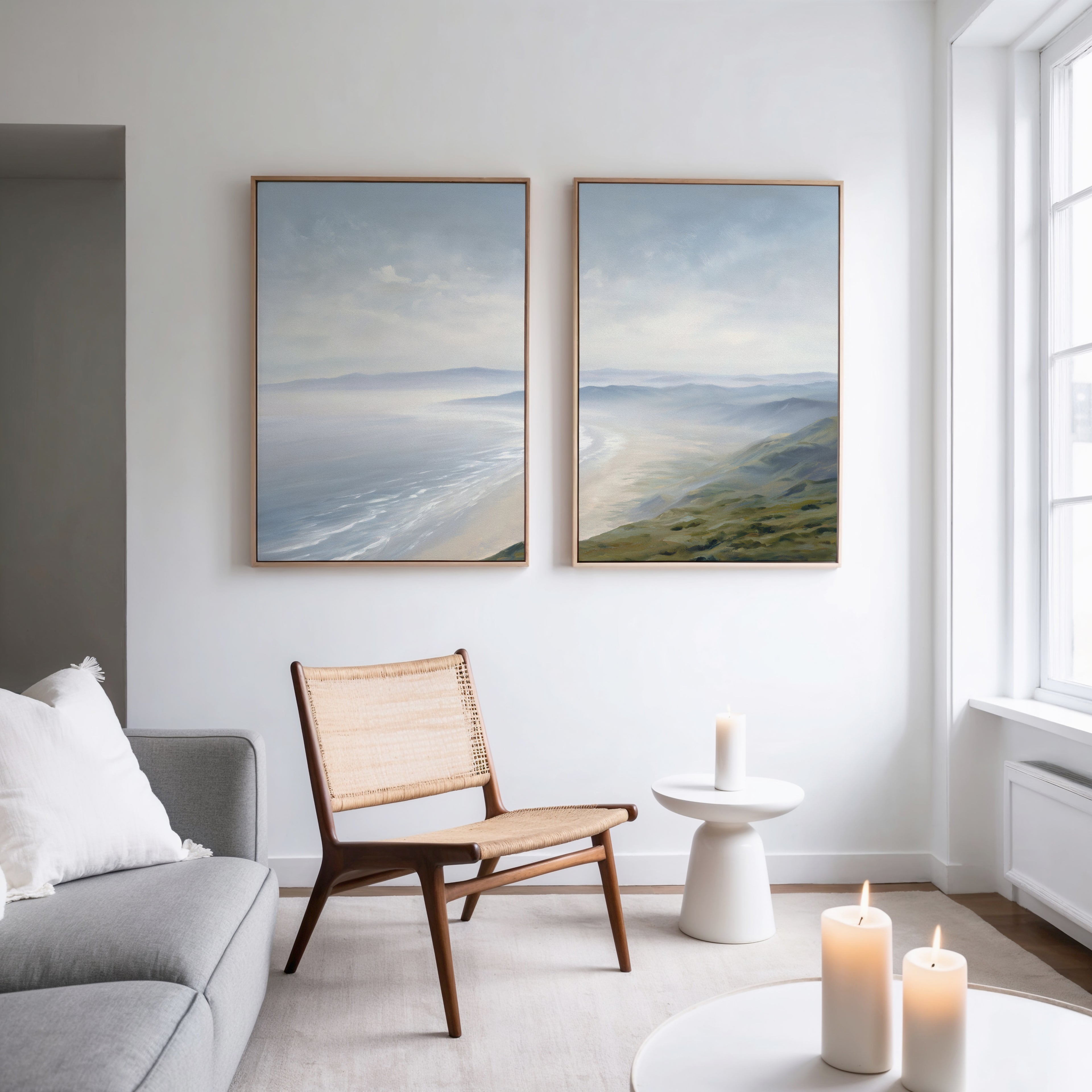 A modern minimal living room features a wooden chair, gray sofa, round white side table, lit candles, and Veiled Shores - Set of 2 coastal landscape canvas artworks hanging on a white wall near a tall window.