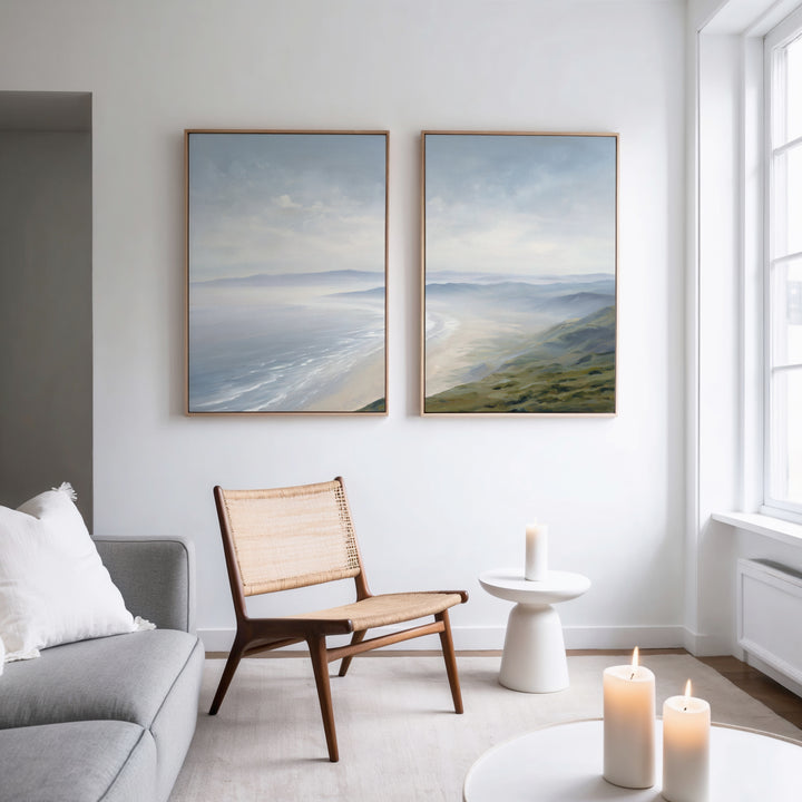 A modern minimal living room features a wooden chair, gray sofa, round white side table, lit candles, and Veiled Shores - Set of 2 coastal landscape canvas artworks hanging on a white wall near a tall window.