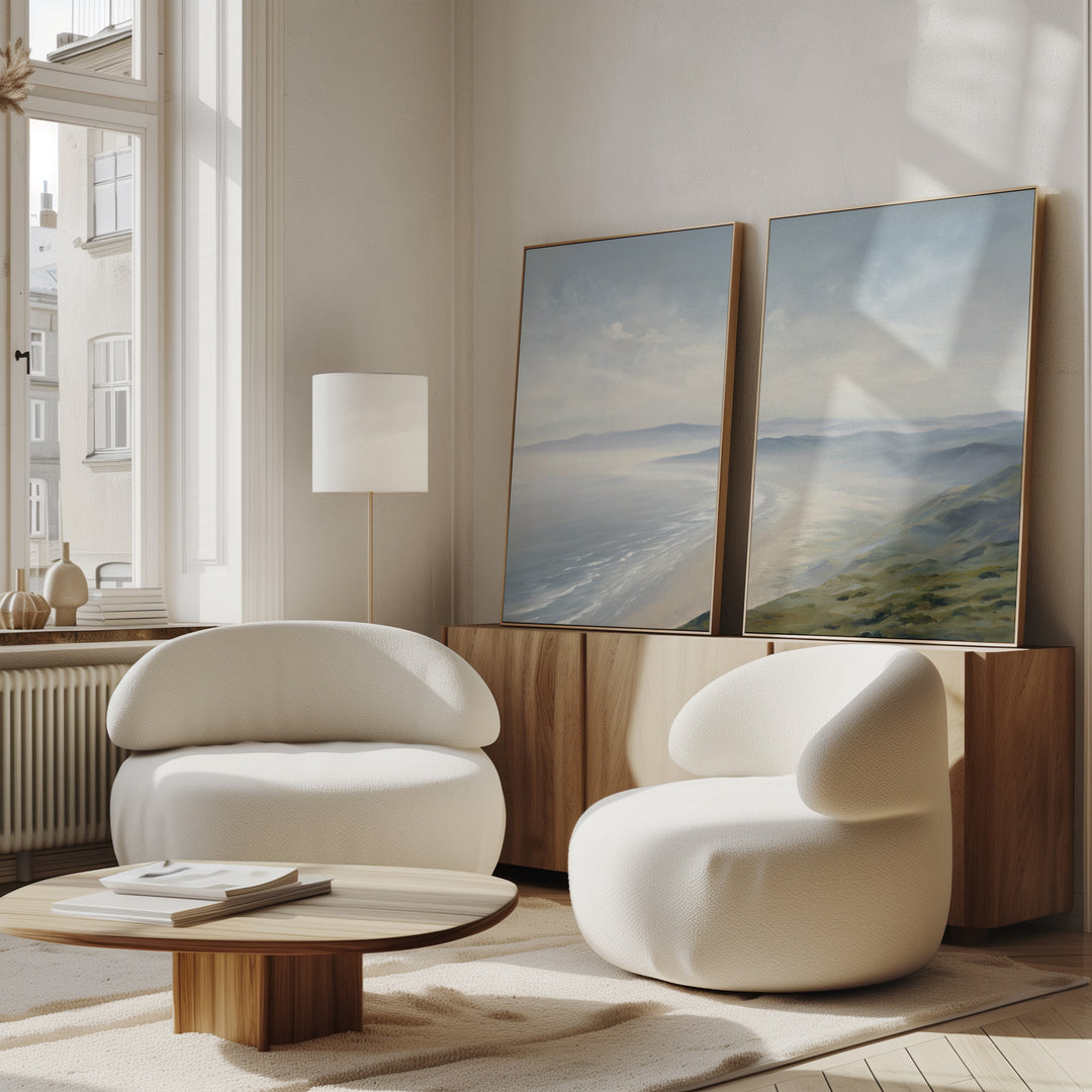 A bright, modern living room with two rounded white chairs, a low wooden coffee table, and Veiled Shores - Set of 2 landscape artworks leaning against a wood sideboard, next to a tall floor lamp by large windows.