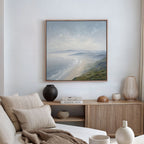 A cozy, minimalist living room with a beige sofa, neutral cushions, and a wooden sideboard topped with vases and books. Above, the Veiled Shores Canvas Art - Square adds tranquility with its serene coastal landscape.