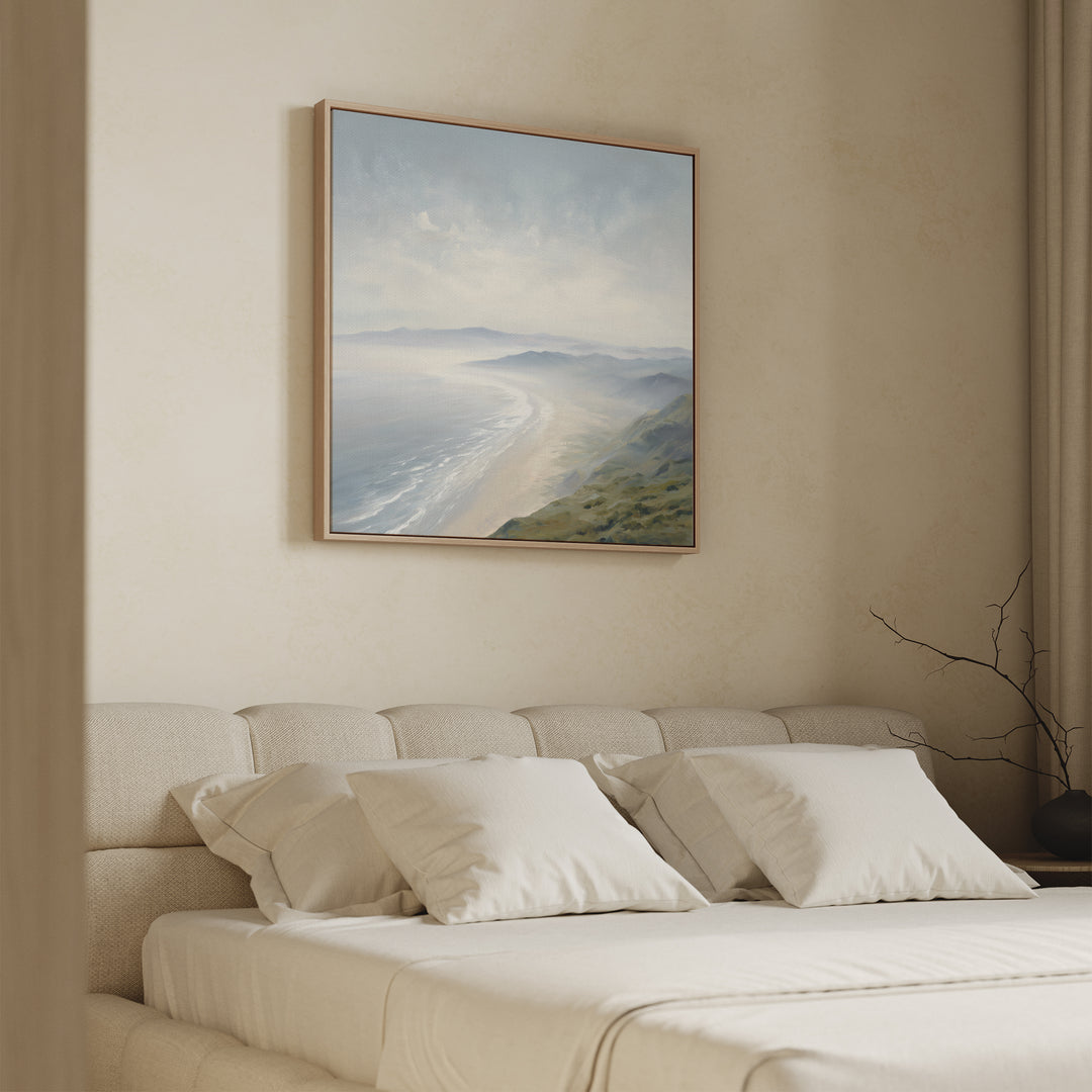 A beige, minimalist bedroom featuring a soft upholstered bed with white pillows. Above the headboard hangs the Veiled Shores Canvas Art - Square, showcasing ocean waves and cliffs. A bare branch decorates the bedside table.