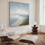 A modern, minimalist room features a white lounge chair and ottoman, wooden flooring, a small wooden side table with a white lamp, and Veiled Shores Canvas Art - Square as an eye-catching coastal landscape on the wall.