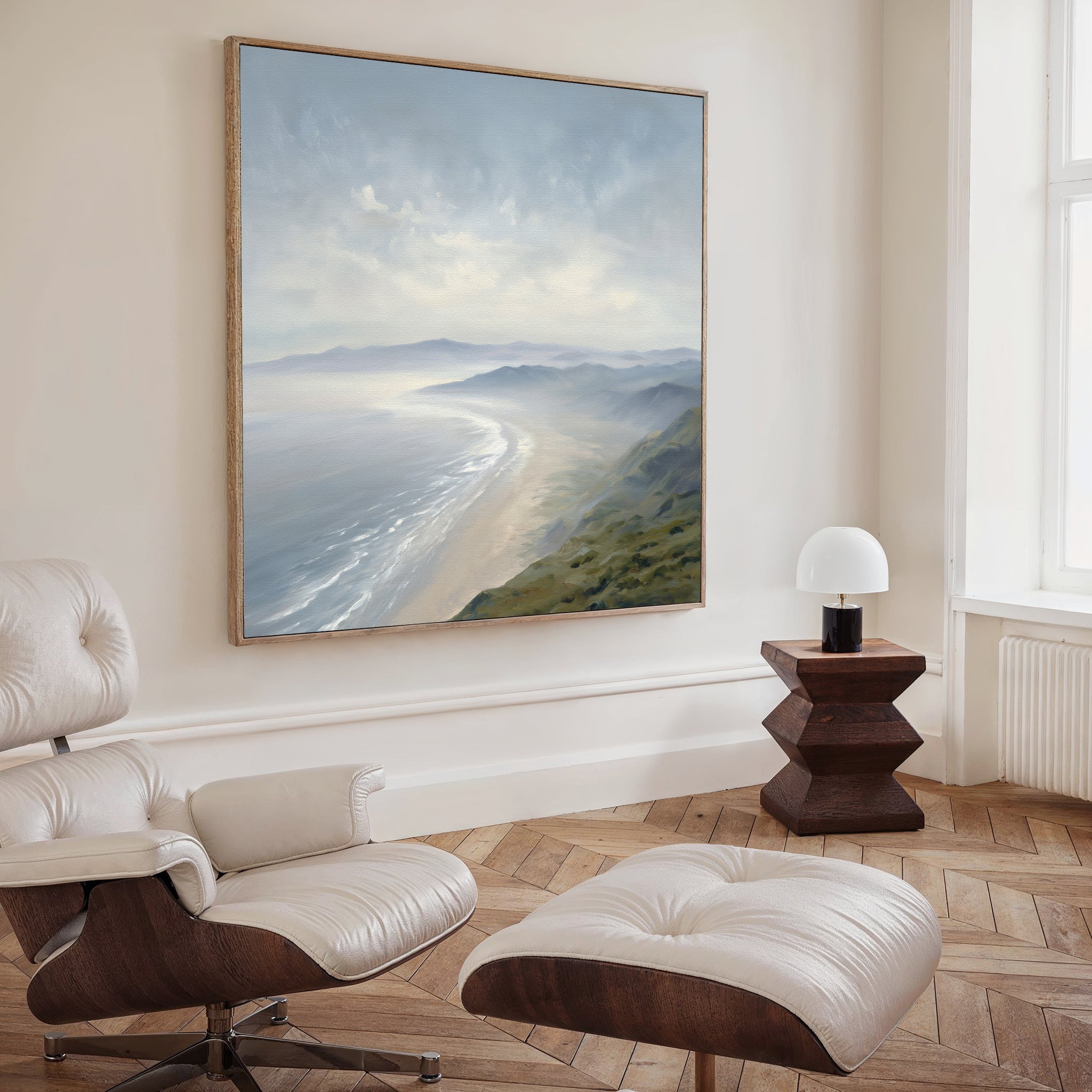 A modern, minimalist room features a white lounge chair and ottoman, wooden flooring, a small wooden side table with a white lamp, and Veiled Shores Canvas Art - Square as an eye-catching coastal landscape on the wall.