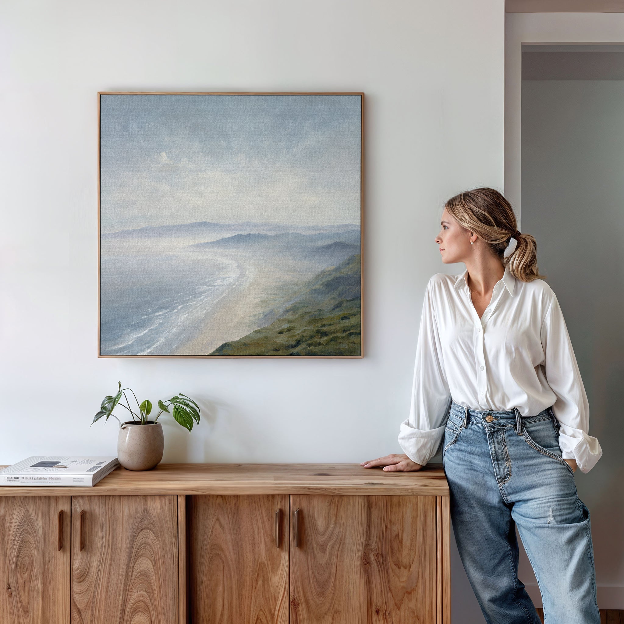 A woman in a white blouse and jeans leans on a wooden cabinet, gazing at the Veiled Shores Canvas Art - Square displayed above. A plant and book decorate the cabinet in this minimalist, bright room.