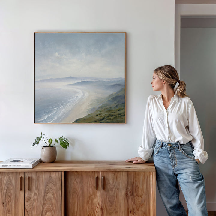 A woman in a white blouse and jeans leans on a wooden cabinet, gazing at the Veiled Shores Canvas Art - Square displayed above. A plant and book decorate the cabinet in this minimalist, bright room.