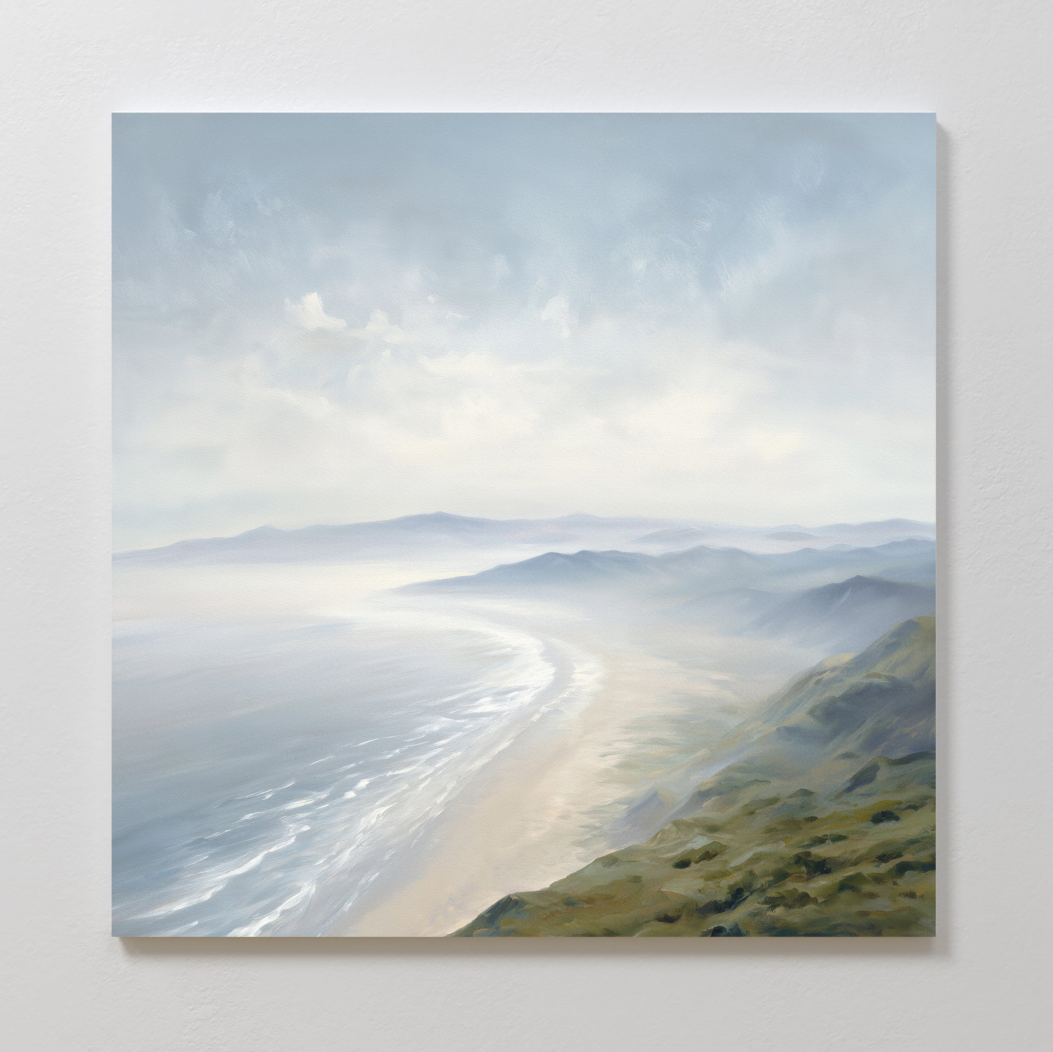 The "Veiled Shores Canvas Art - Square" showcases a misty coastal landscape with rolling hills and a sandy shoreline, soft blue-gray skies, and gentle waves, creating a peaceful atmosphere—ideal as coastal wall art.