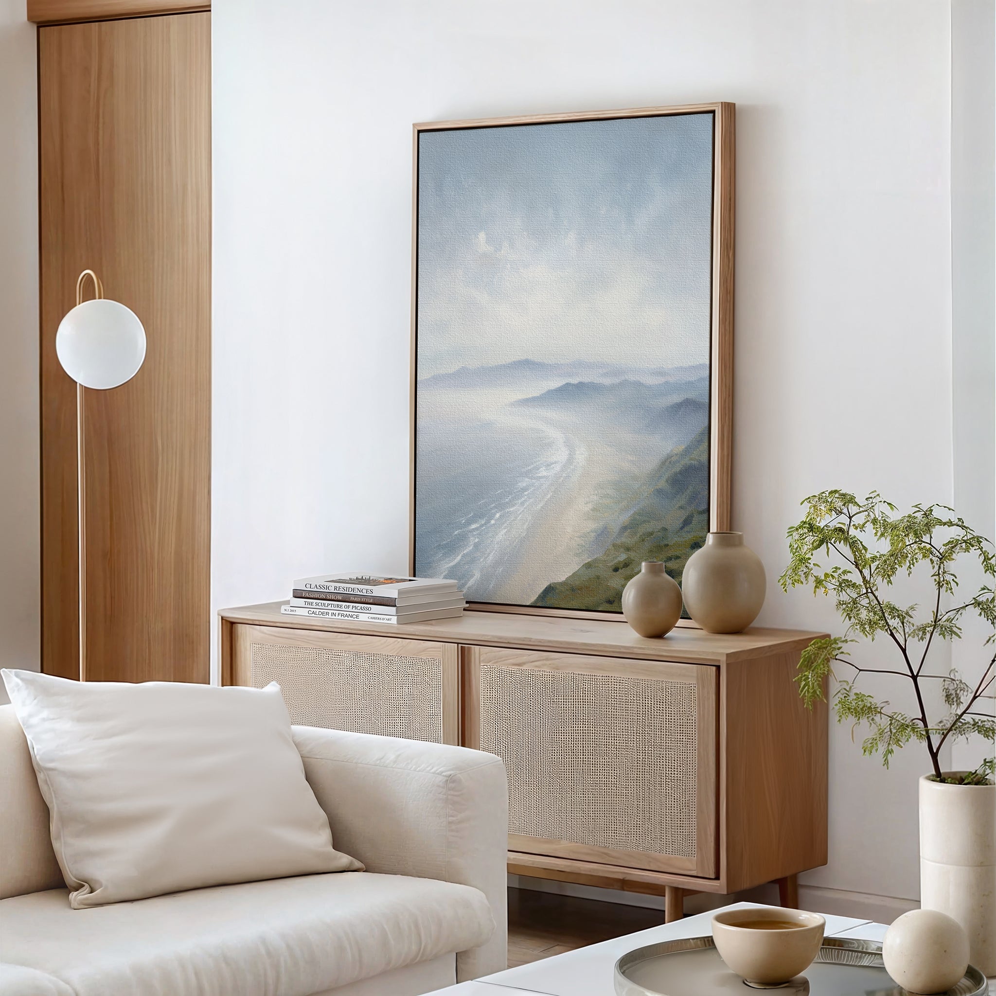 A modern, minimalist living room with a light wood sideboard, Veiled Shores Canvas Art - Vertical above, ceramic vases, books, a white sofa with pillows, and a potted plant on a white table.