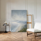 The Veiled Shores Canvas Art - Vertical leans against a white paneled wall near an empty frame. Nearby, a cushioned chair and round side table with candles on herringbone flooring enhance the tranquil, serene setting.
