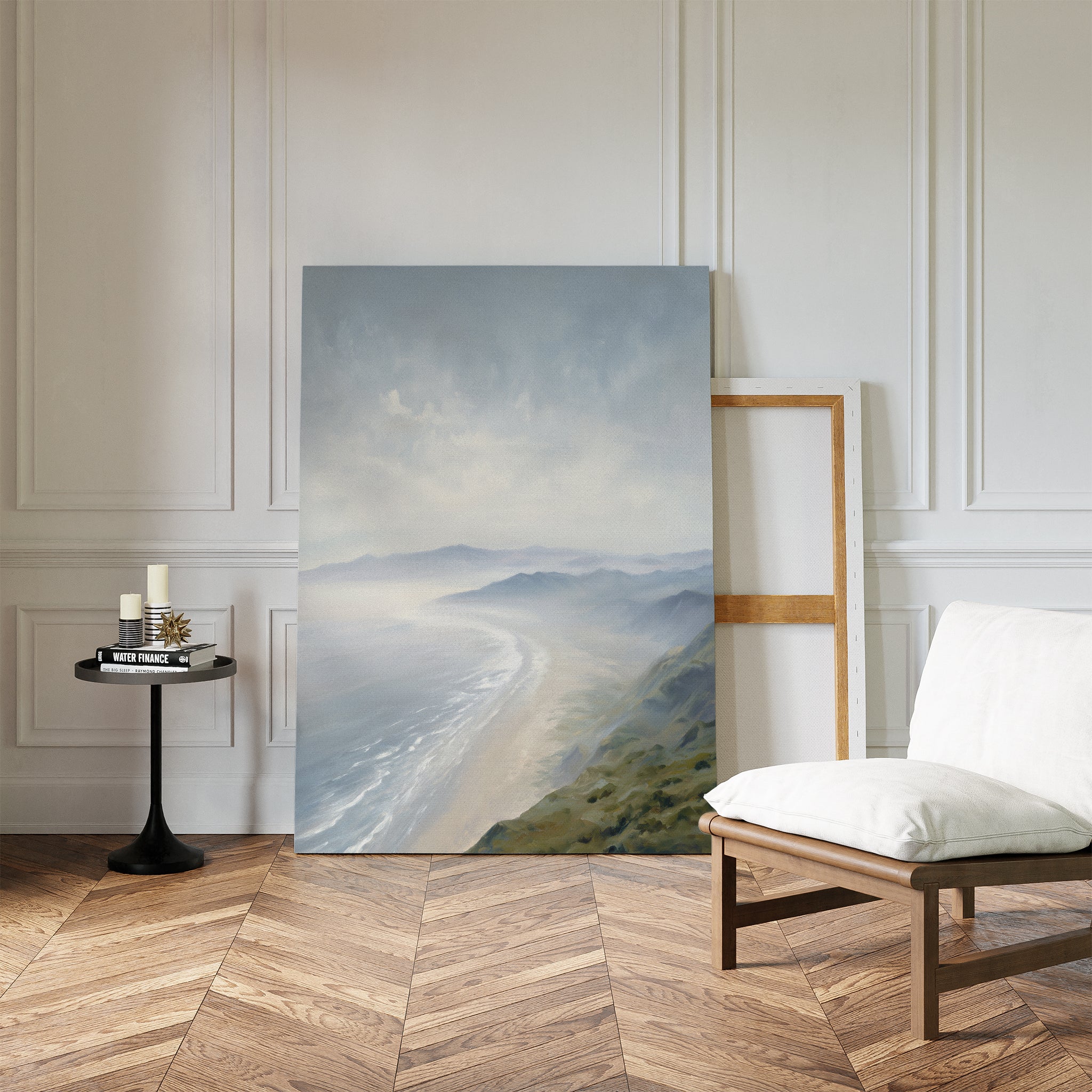 The Veiled Shores Canvas Art - Vertical leans against a white paneled wall near an empty frame. Nearby, a cushioned chair and round side table with candles on herringbone flooring enhance the tranquil, serene setting.