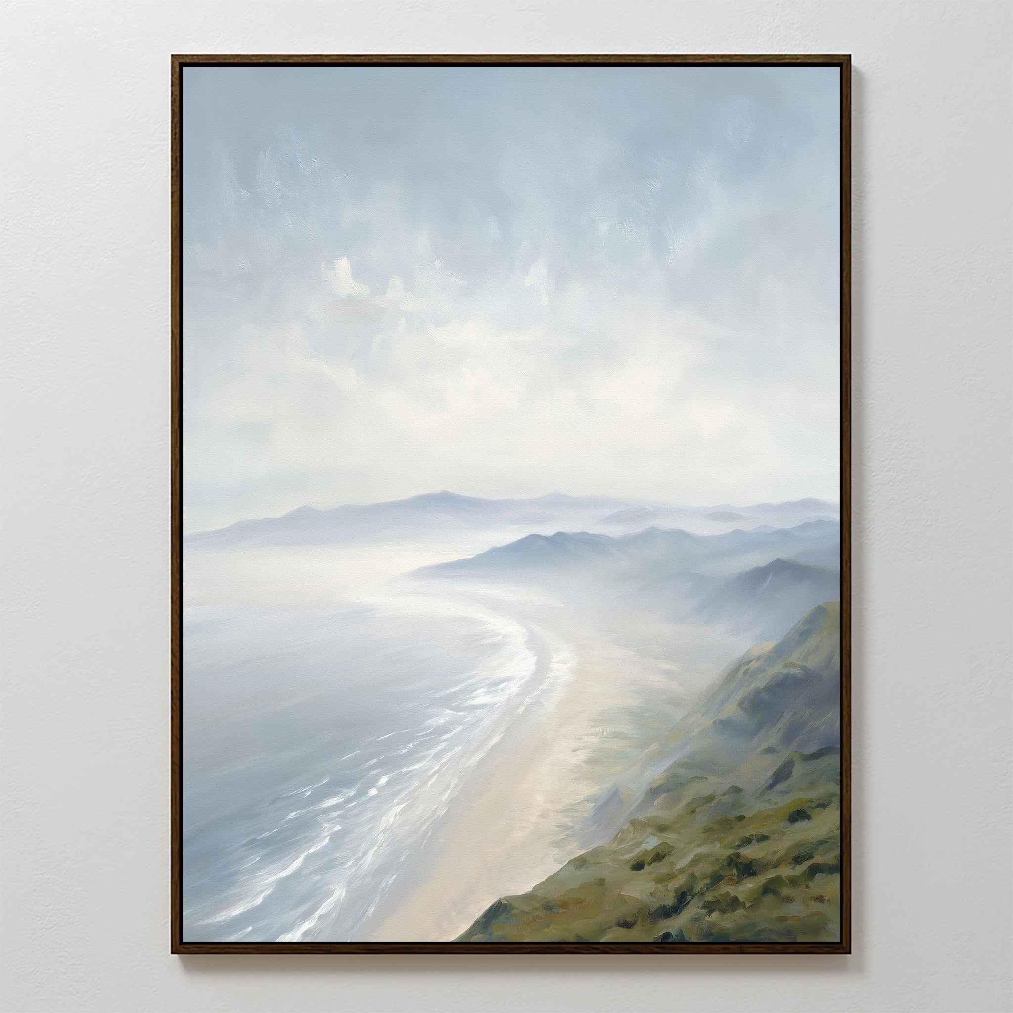 The Veiled Shores Canvas Art - Vertical features a tranquil, hazy coastline with gentle waves, sandy beach, and green hills beneath a cloudy sky. This framed coastal landscape is displayed on a white wall from an elevated perspective.