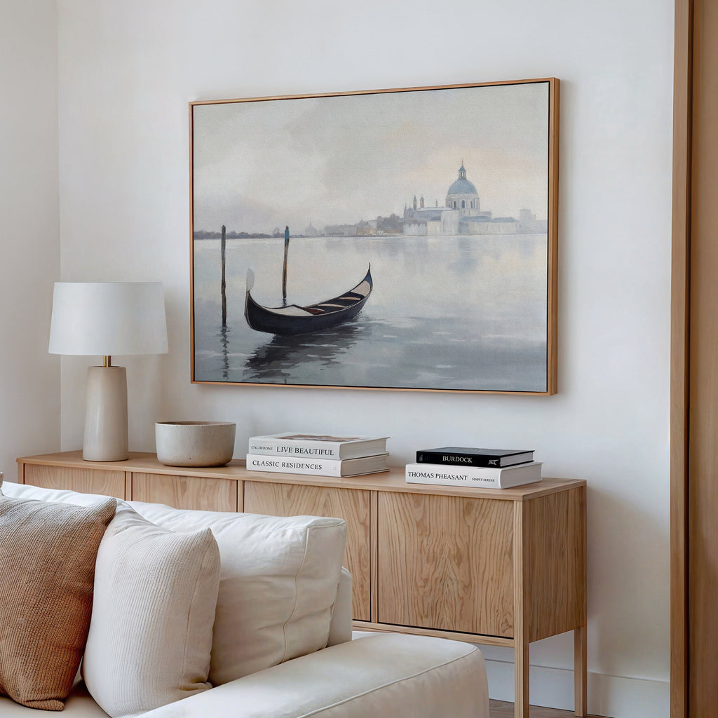 A minimalist living room features a wooden sideboard with books, a lamp, and a bowl. Above it hangs the Venetian Stillness Canvas Art - Horizontal, depicting a gondola on calm water with a domed building in the distance.