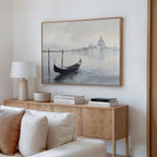 A minimalist living room features a wooden sideboard with books, a lamp, and a bowl. Above it hangs the Venetian Stillness Canvas Art - Horizontal, depicting a gondola on calm water with a domed building in the distance.