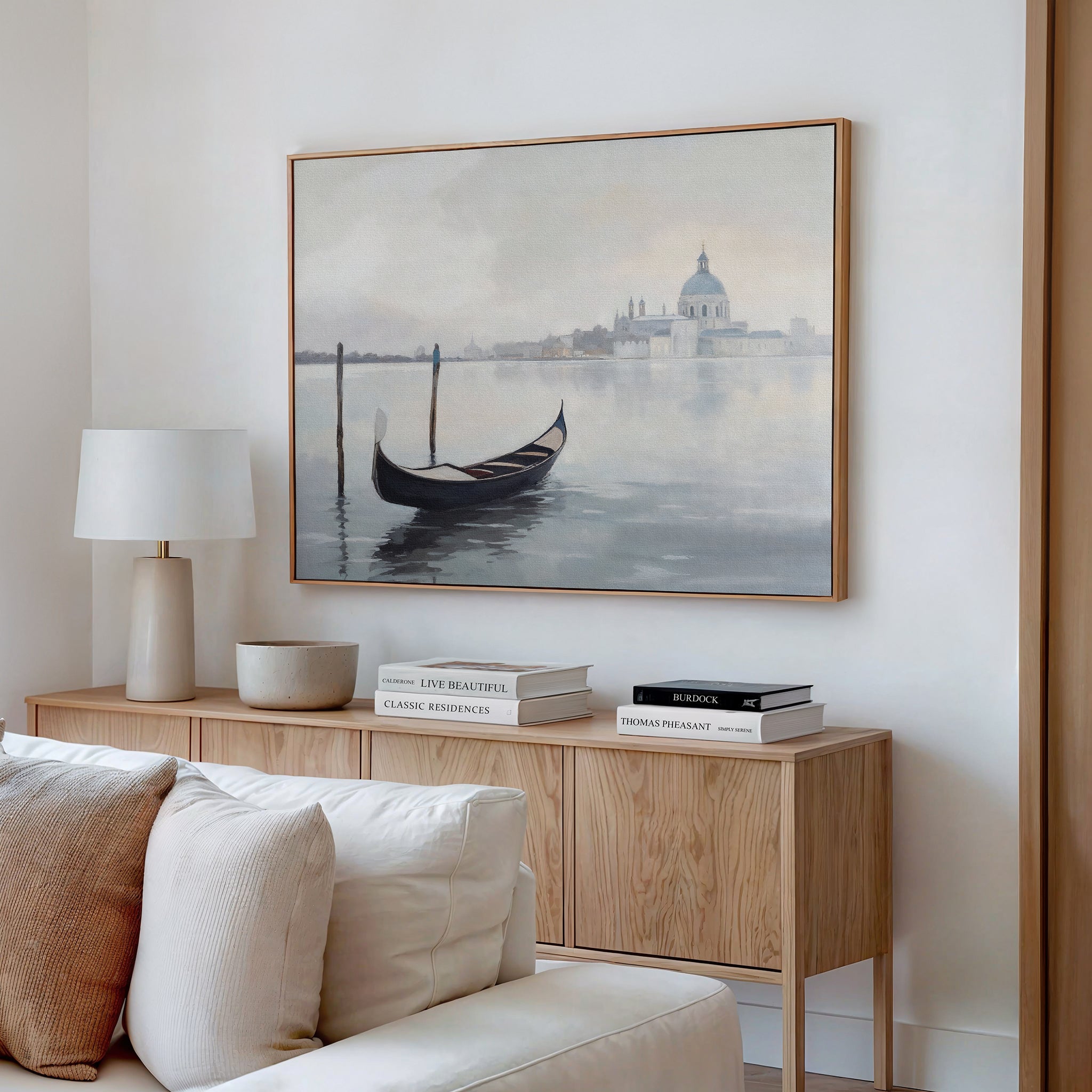 A minimalist living room features a wooden sideboard with books, a lamp, and a bowl. Above it hangs the Venetian Stillness Canvas Art - Horizontal, depicting a gondola on calm water with a domed building in the distance.