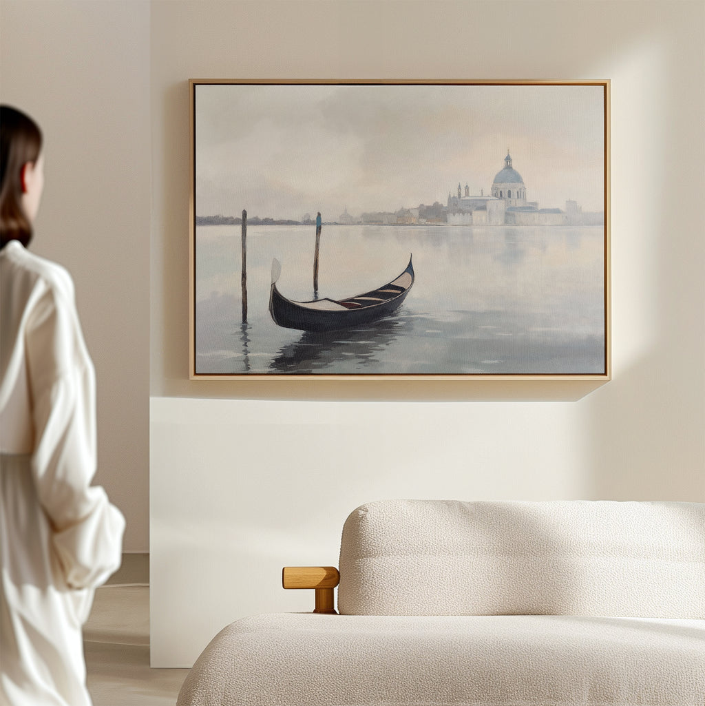 A woman stands in a minimal, light room, gazing at the Venetian Stillness Canvas Art - Horizontal featuring a gondola on calm water with a distant city and domed building, displayed above a beige sofa.
