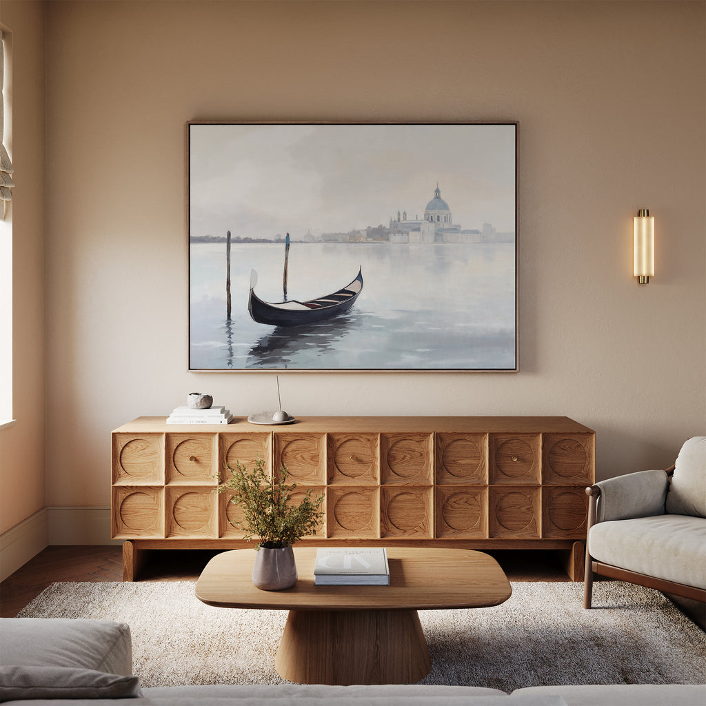 A modern living room with neutral walls, a wooden sideboard, coffee table, sofa, and the Venetian Stillness Canvas Art - Horizontal featuring a gondola on the water with buildings in the background above the sideboard.