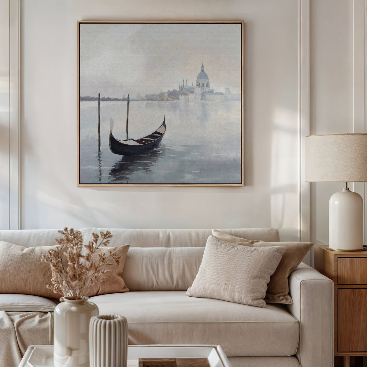 A modern living room features a beige sofa, neutral cushions, a side table with a lamp, a vase of dried flowers, and the "Venetian Stillness Canvas Art - Square," lending timeless European charm to the serene space.