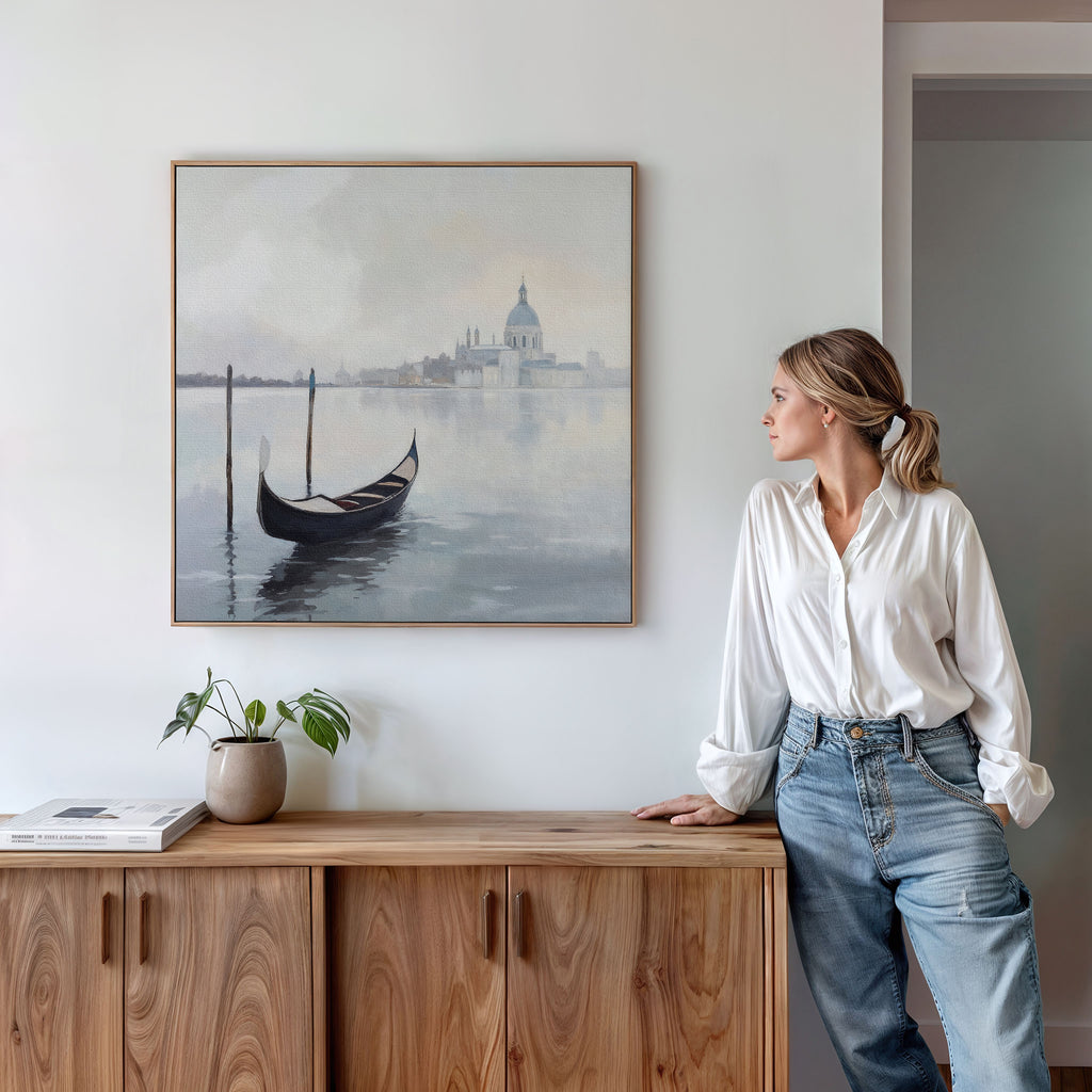A woman in a white blouse and jeans leans against a wooden cabinet, gazing at the "Venetian Stillness Canvas Art - Square," which captures European charm with its gondola and cityscape scene displayed above the cabinet.