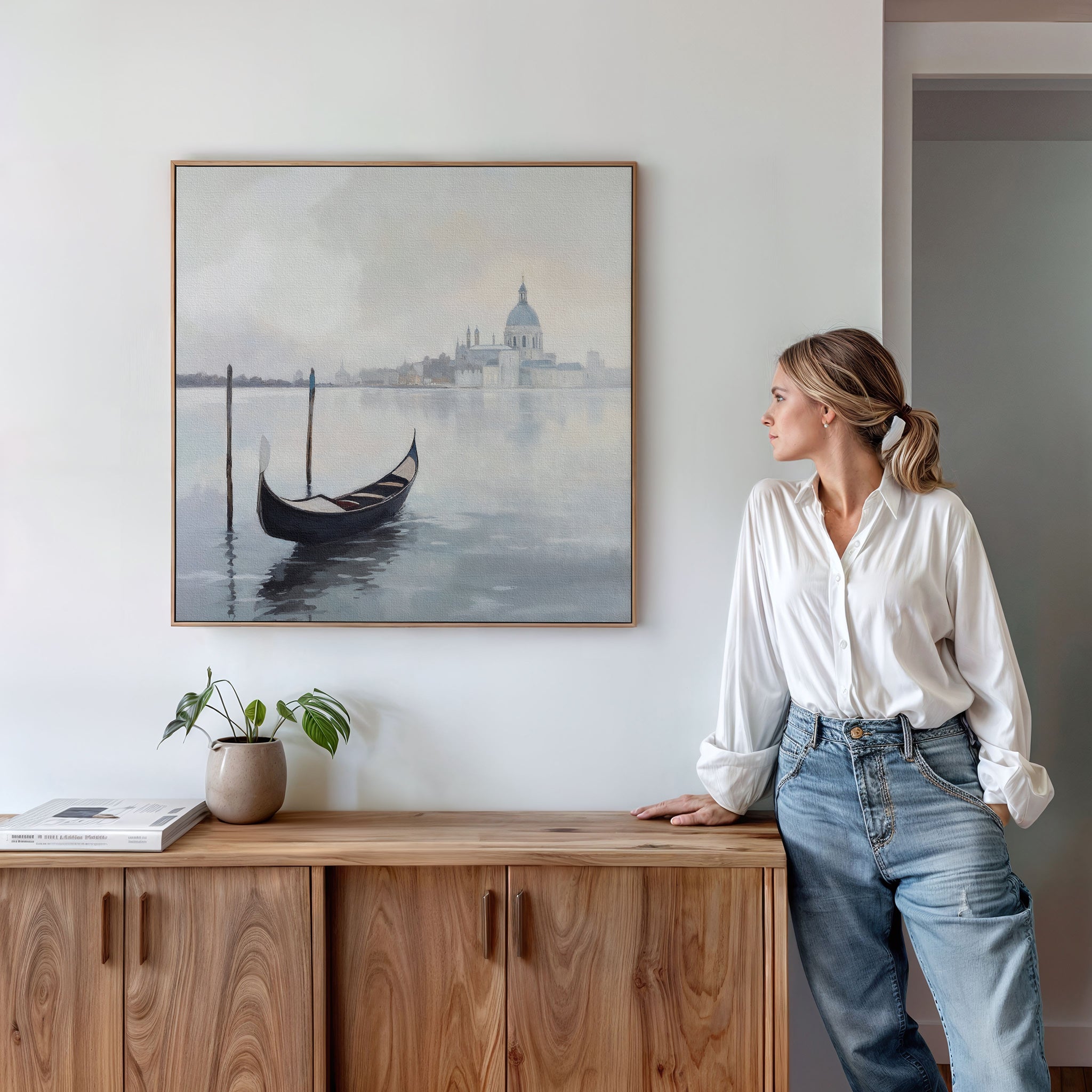 A woman in a white blouse and jeans leans against a wooden cabinet, gazing at the "Venetian Stillness Canvas Art - Square," which captures European charm with its gondola and cityscape scene displayed above the cabinet.